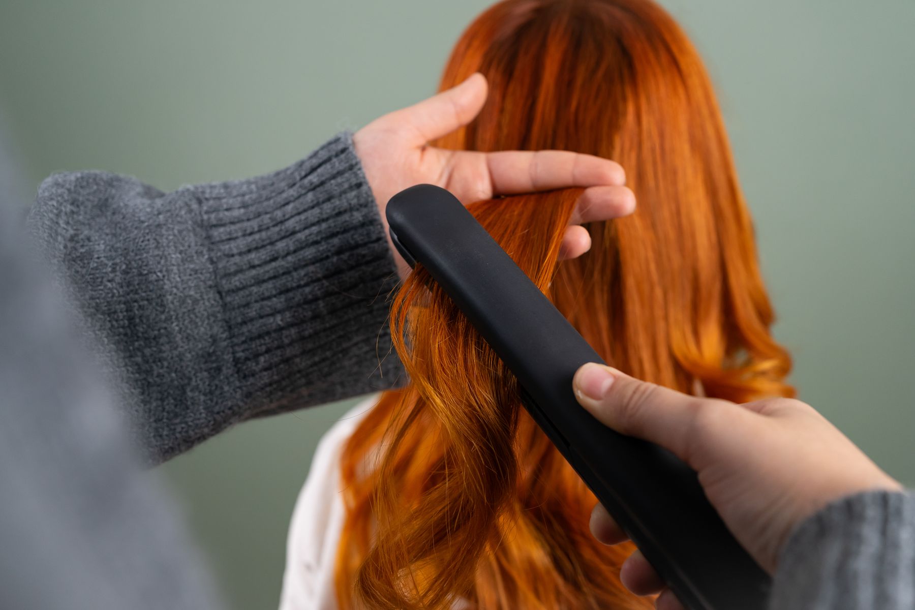 Hands styling long red hair with a flat iron